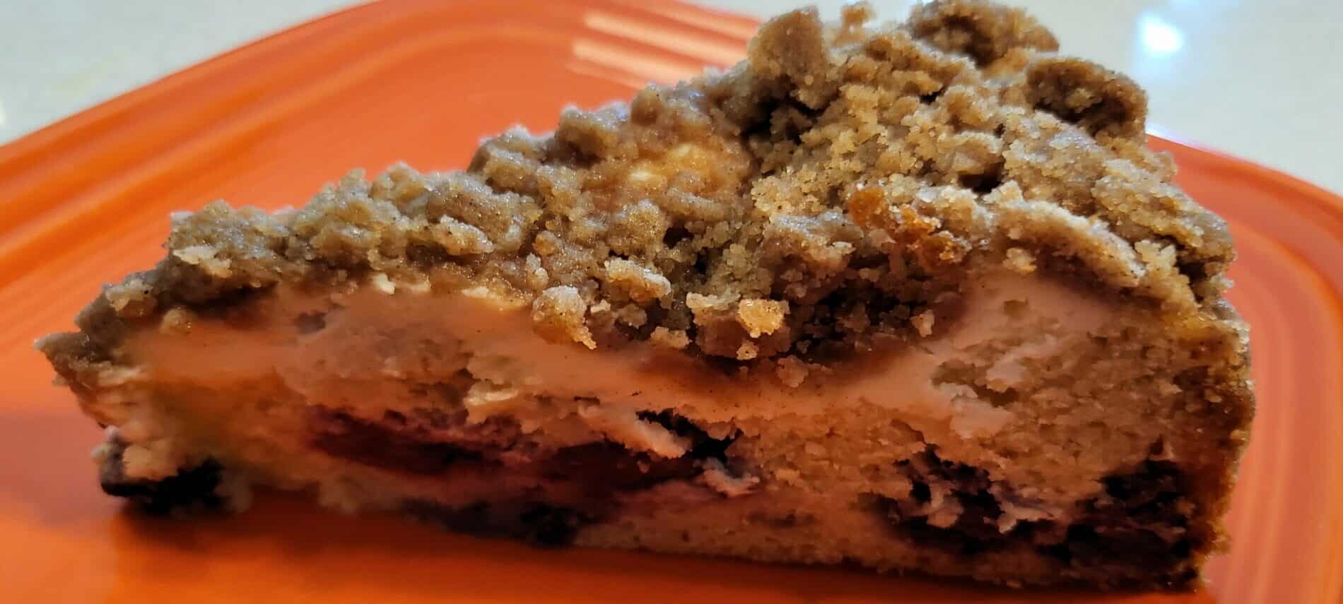 slice of coffee cakes with a crumble topping, a cream cheese layer and cake layer with blackberries on orange plate
