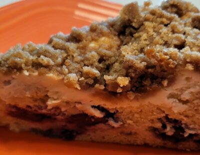 slice of coffee cakes with a crumble topping, a cream cheese layer and cake layer with blackberries on orange plate
