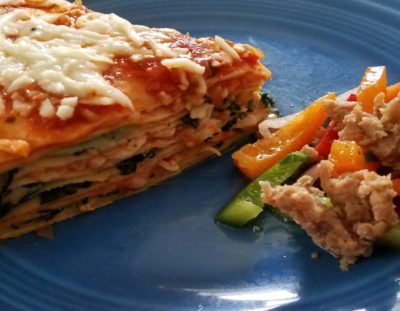 Triangle piece of lasagna on blue plate with red, orange and green pepper and sausage salad