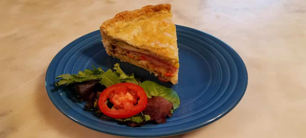 layers of ham, salami, cheese and olive salad in pie crust on blue plate with greens and tomato slice