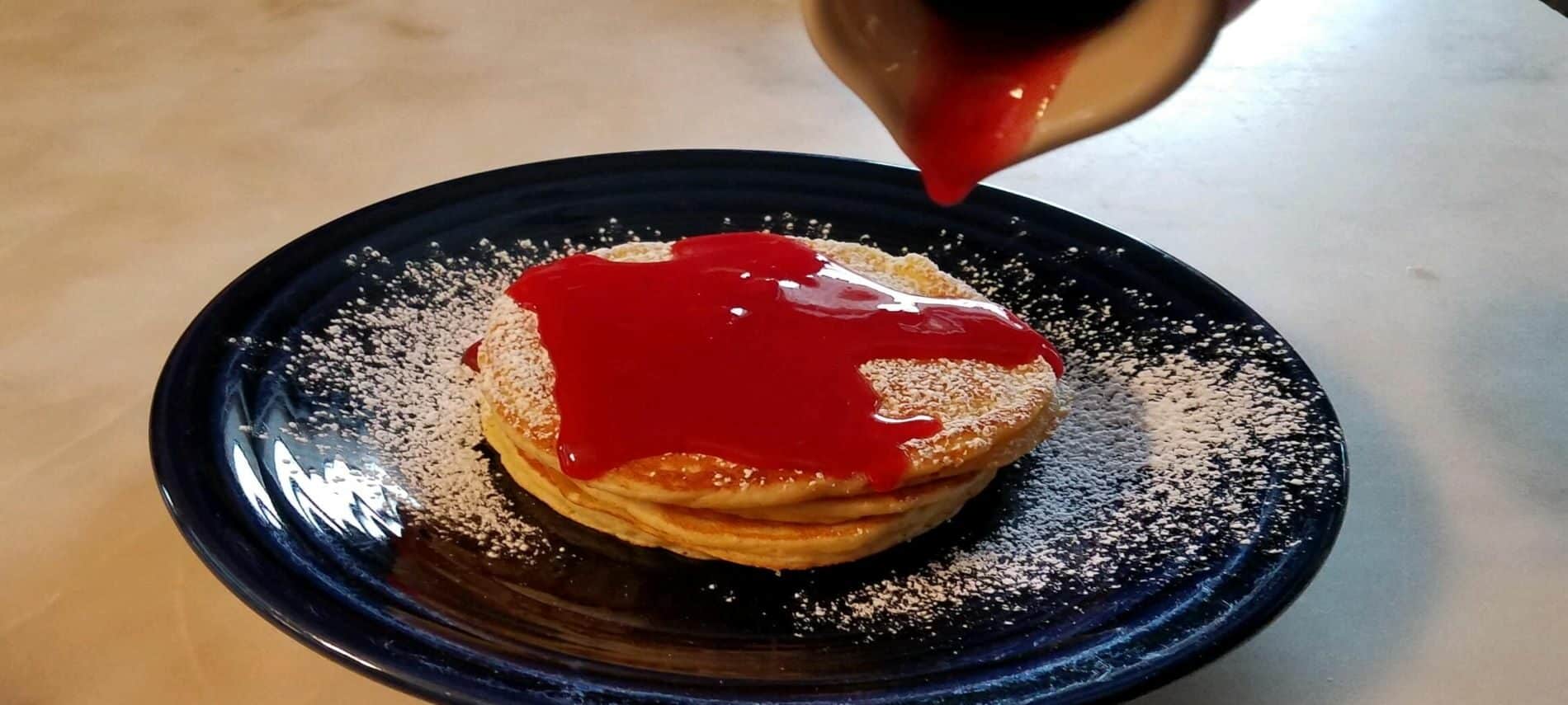 dark blue plate with golden brown pancakes with cranberry syrup poured from pitcher on top of pancakes