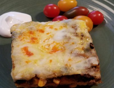 Layered South of the Border Breakfast Lasagna with melted cheese on top, served on a green plate with sour cream and multi colored cherry tomatoes