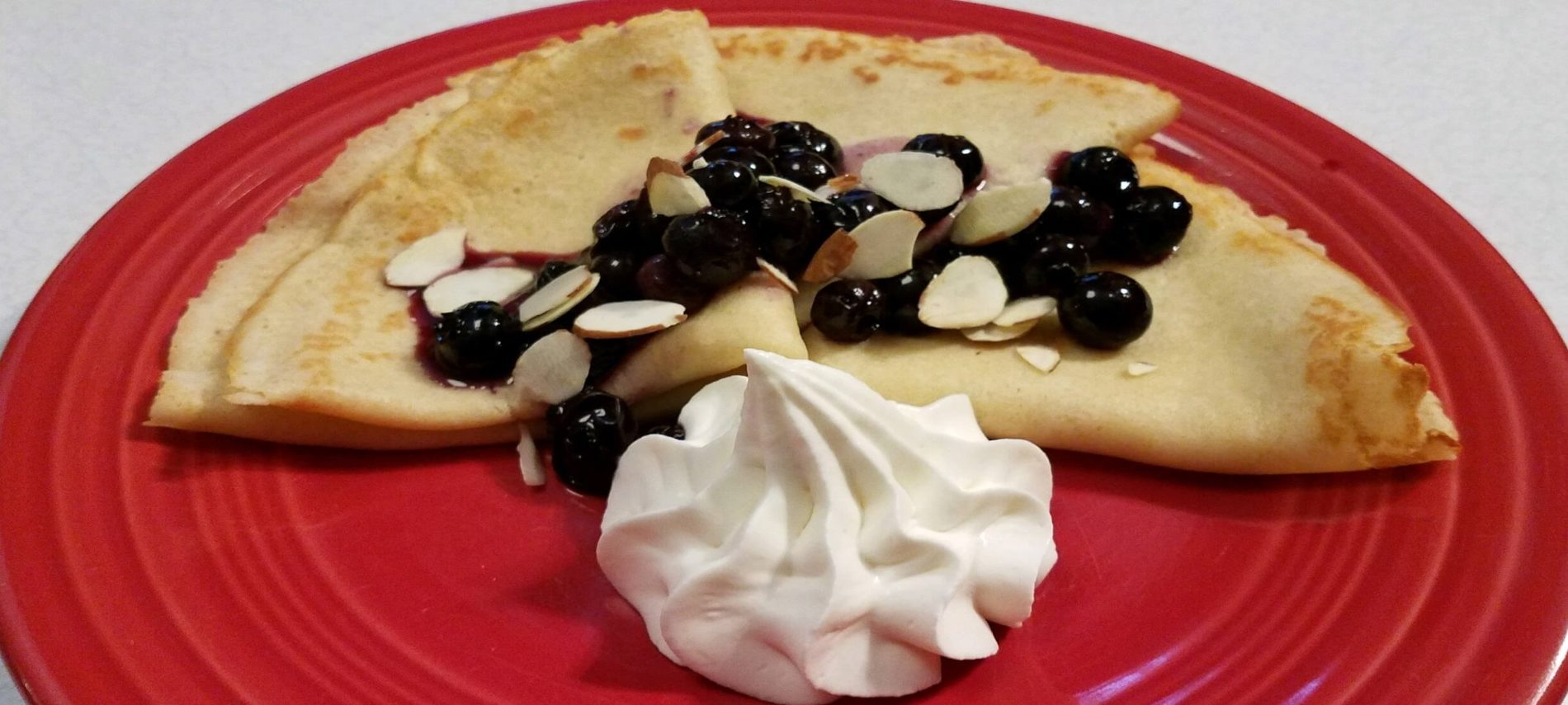Three swedish pancakes lightly browned folded into triangles, topped with blueberries and side of whipped cream on a red plate