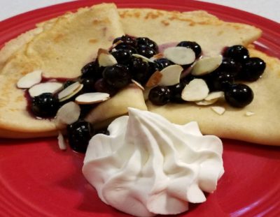 Three swedish pancakes lightly browned folded into triangles, topped with blueberries and side of whipped cream on a red plate