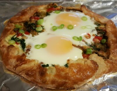 On a foil lined pan is a rustic pie with sausage, peppers, cheese, and baked egg, topped with green onions