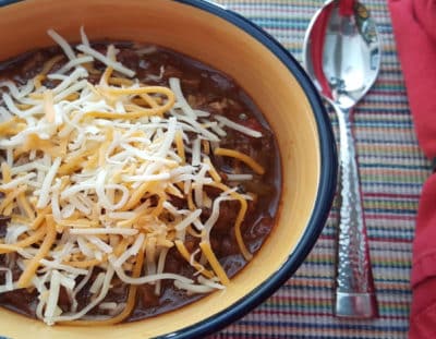 yellow and blue bowl of chili with grated cheddar and jack cheese with spoon and red napkin with fleur de lis napkin holder