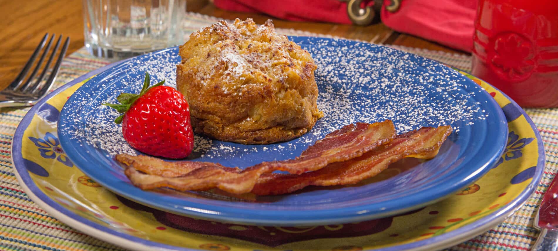Blue ceramic plate on a colorful charger topped with bacon, red strawberry and golden baked french toast