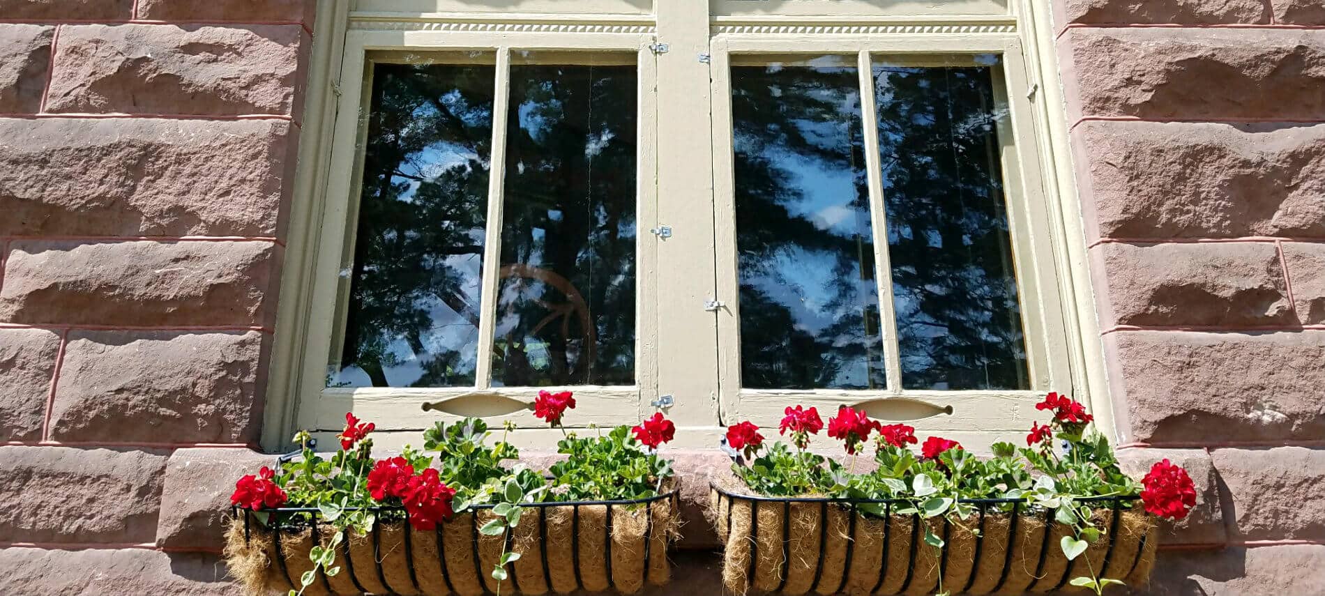 Close up view of exterior window surrounded by reddish stone and two window boxes with bright red flowers