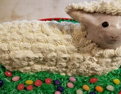 Cake in the form of a lamb with swirled white buttercream, glazed face, on a red plate covered with green shredded coconut and assorted colors of jelly beans