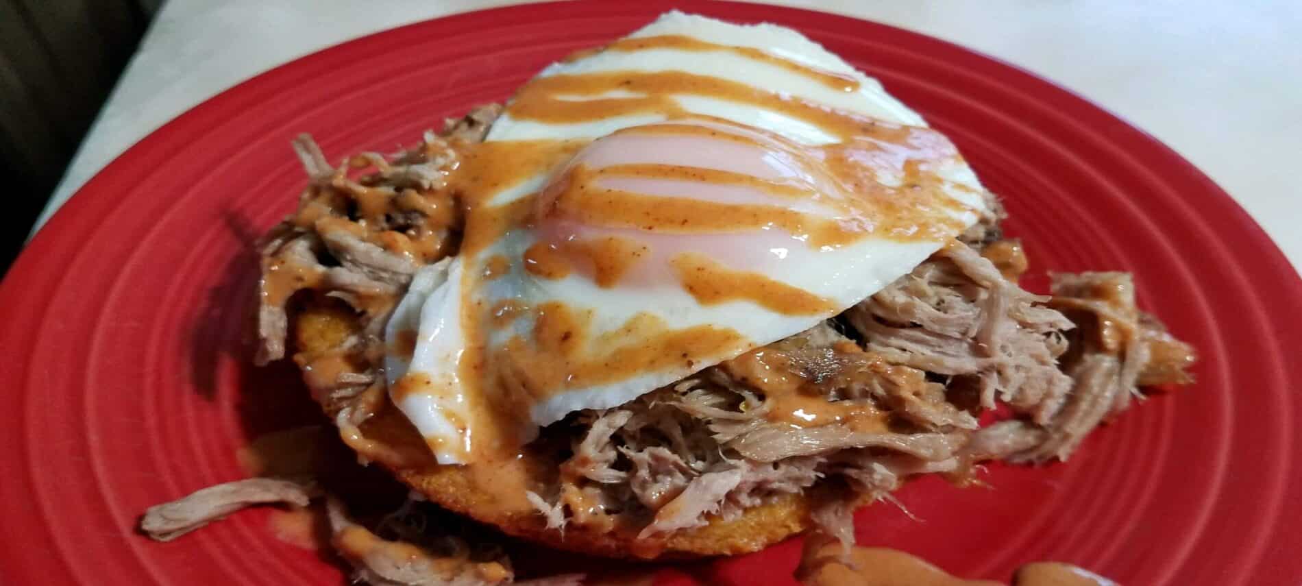 crispy cornbread covered with pulled pork, topped with over easy egg, drizzled with a light red sauce on a red plate
