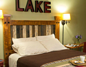 mission style wooden headboard with wood block letters on wall above spelling out the word lake against soft green wall with shaded sconce next to bed covered in neutral brown spread and four pillows with crip white pillow cases.