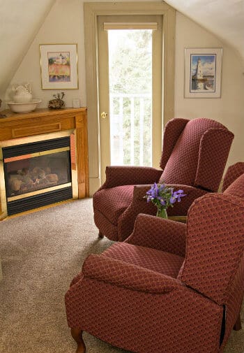 Beige carpeted room with sloped ceilings, glass door, two burgundy upholstered wingback chairs and corner fireplace