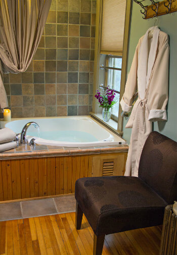 Bathroom with wood floors, upholstered chair, soaker tub with tiled wall, and window with fresh flowers
