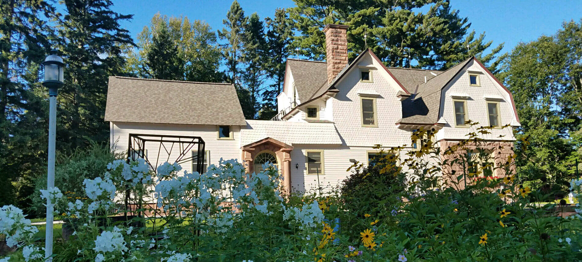 Exterior view of inn surrounded by tall green trees, colorful flowers and blue skies
