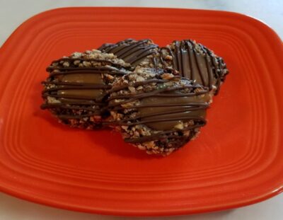 four chocolate cookies coated in pecans with caramel in the center and drizzled with chocolate on an orange plate