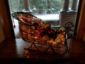 wicker tabletop sleigh with white lights filled with red and gold Christmas ornaments on wood table in front of large window